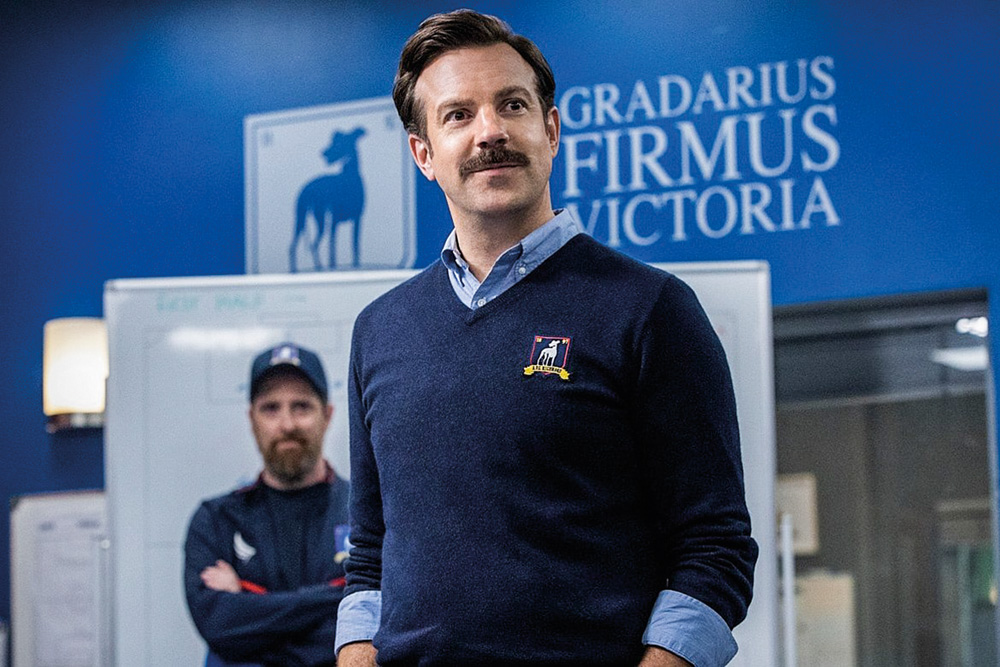 A imagem mostra o ator Jason Sudeikis, na série Ted Lasso, com um suéter com bordado do clube no qual é treinador e ele olhando para o vestiário onde está seu time