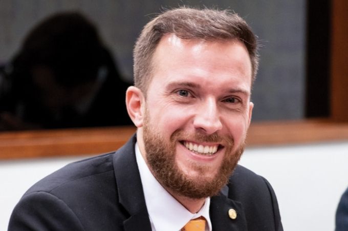 vinicius poit está de terno, em uma sala, sorrindo