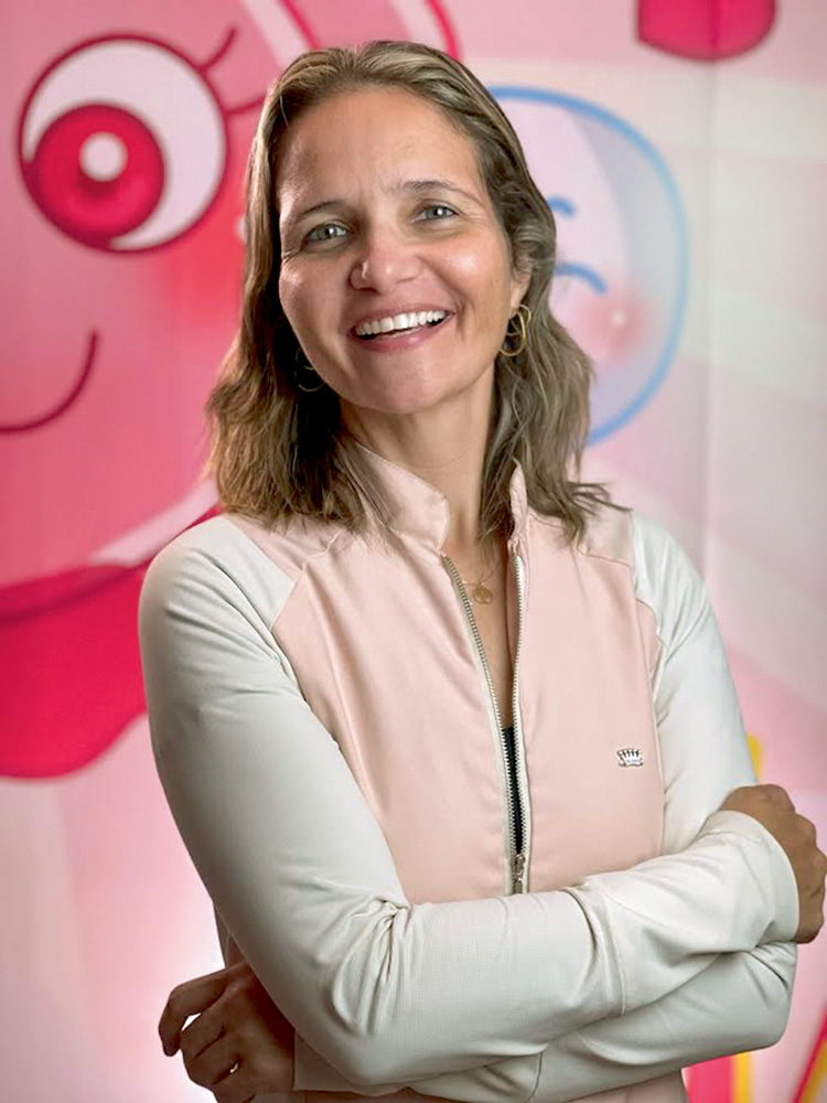 Letícia Piccolo A imagem mostra Letícia, de braços cruzados, sorrindo para a câmera em pé.