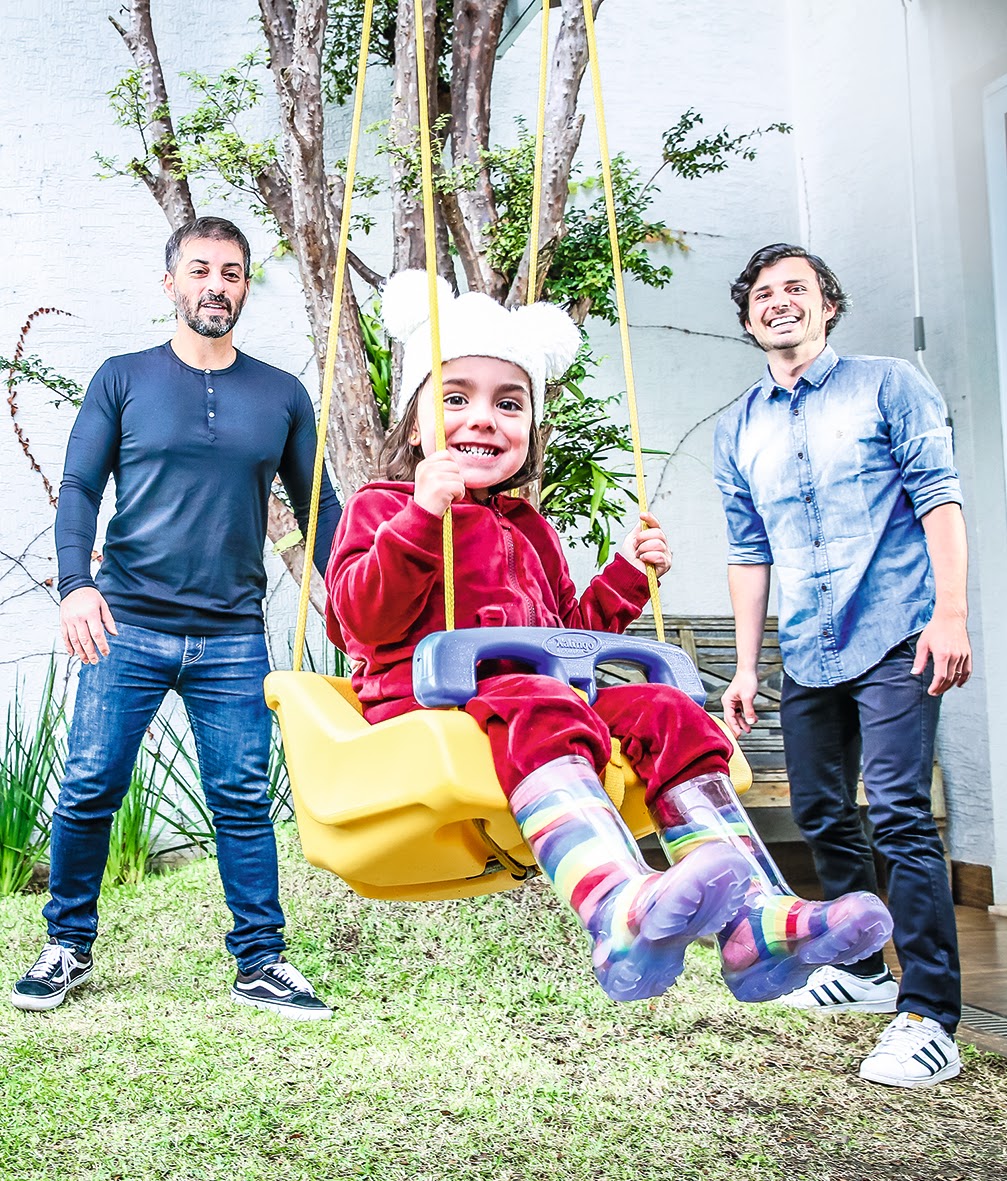 Rogerio Capela e Gustavo França com a filha Alice 2 A imagem mostra Rogerio e Gustavo e a filha Alice em um balanço. Os três estão sorrindo.