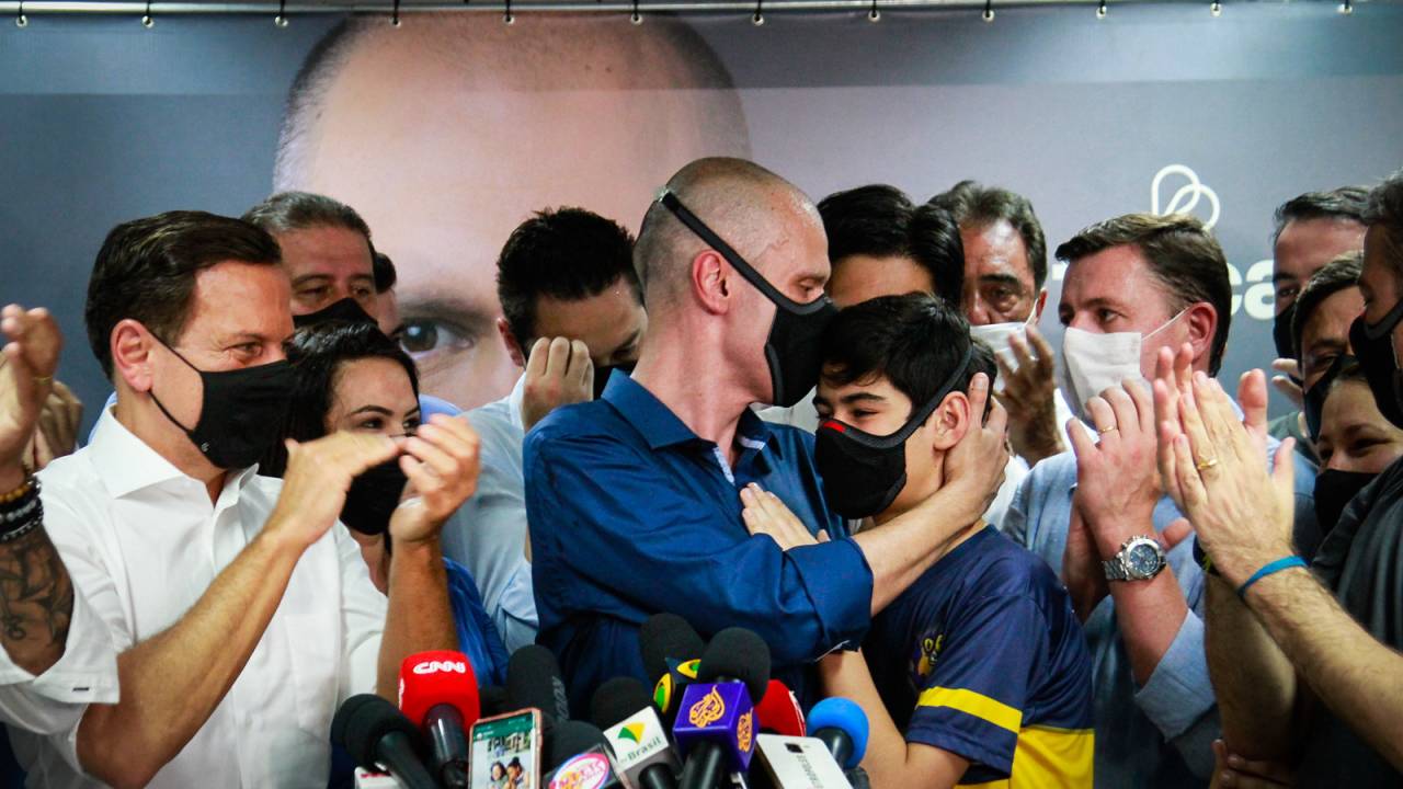 Foto mostra Bruno beijando a testa de Tomás, ao redor, governador João Doria e outros presentes aplaudem o momento. Em uma bancada na frente de Bruno e Tomás, dezenas de microfones de emissoras de TV e rádio