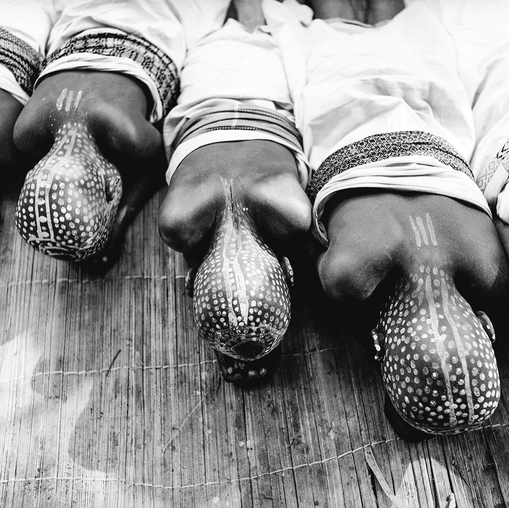 A imagem, em preto e branco, mostra três pessoas negras, com a cabeça inteira pintada, deitadas no chão. Ela estão com a cara virada para o chão, então é visto suas nucas e a pintura nelas.