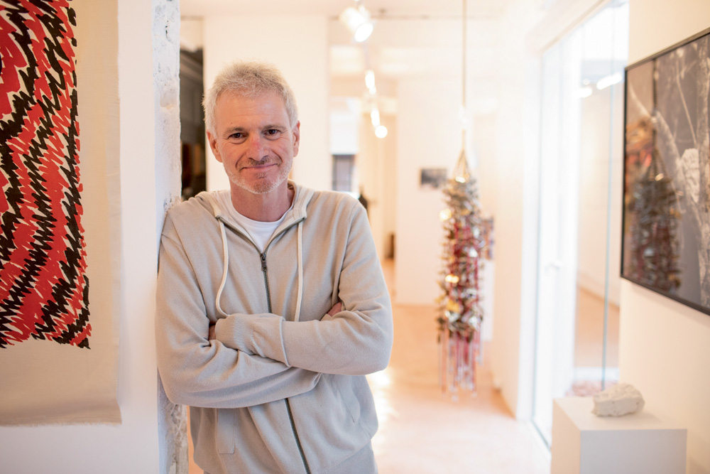Homem grisalho se apoia em uma parede de uma galeria com os braços cruzados e sorri para a foto