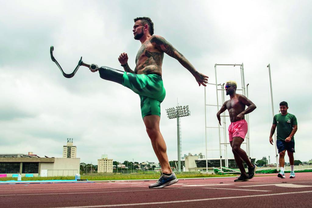 Atleta Vinícius Rodrigues, que tem uma perna amputada, levantando-a na pista de corrida. Há outros dois atletas atrás dele