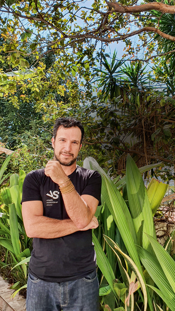 André Palhano, fundador da Virada Sustentável, posa de braços cruzados, uma das mãos no queixo, vestindo camiseta preta. Ao fundo, árvores e arbustos.