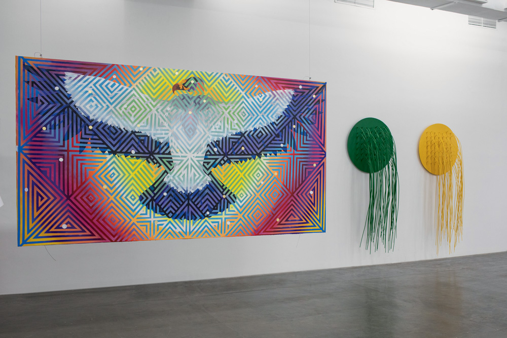 BIENAL A imagem mostra uma obra de Daiara. É um grande gavião, com a cabeça colorida. Ele está de asas abertas, em um único plano. O fundo é colorido, com ele sobre o que parece um sol. Há também linhas e contornos sobre toda obra.
