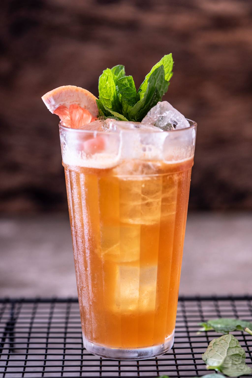 Drinque de cor alaranjada em copo tipo americano grande coberto por hortelã e fatia de laranja.