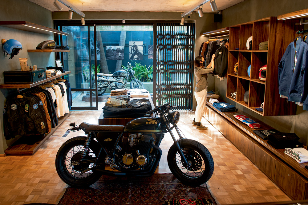 A imagem mostra a fachada da Deus Ex Machina. Nela, há roupas em exibição e uma moto no centro da loja.