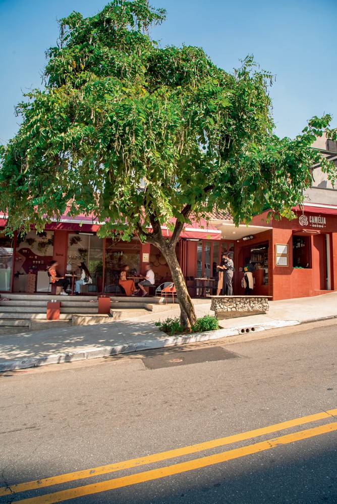 Fachada do Camélia Òdòdó, pintada em cor avermelhada. Na calçada, algumas mesas estão espalhadas. À frente, uma grande árvore faz sombra no ambiente