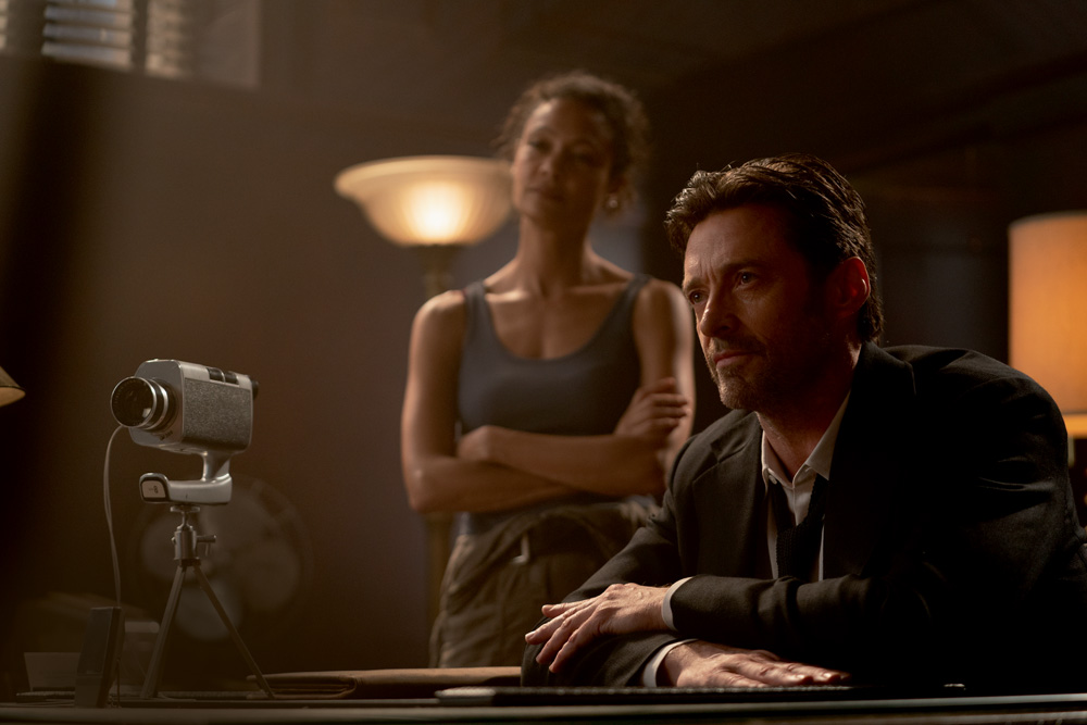 A imagem mostra Hugh Jackman sentado sob uma mesa com uma câmera em sua frente gravando o depoimento de alguém à frente do ator. Ele está com a expressão séria.