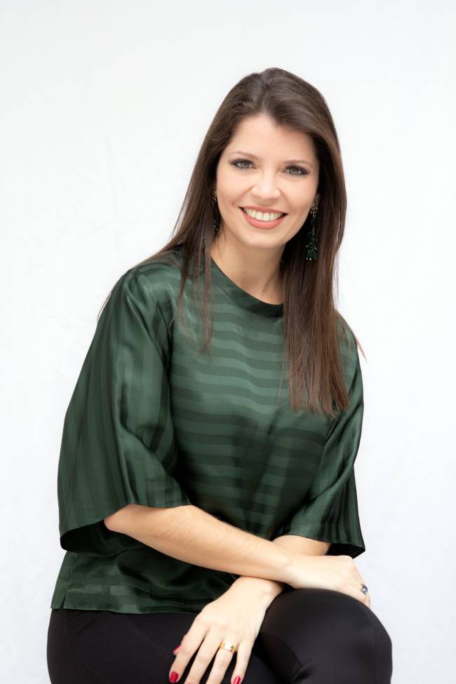 A empreendedora do mercado editorial Caroline Dias sorri para a foto sentada. Veste uma blusa verde-escuro e tem cabelos castanho escuro longos.