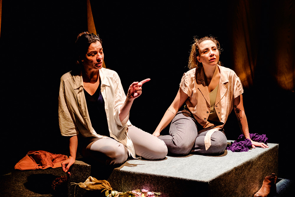 Duas mulheres estão em um palco de teatro. Elas estão em cima de uma mesa, com semblante de dúvida, e estão conversando