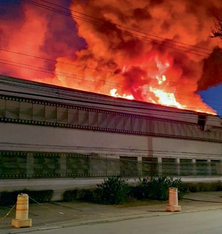 Foto mostra um galpão e, acima, muita fumaça e fogo no céu