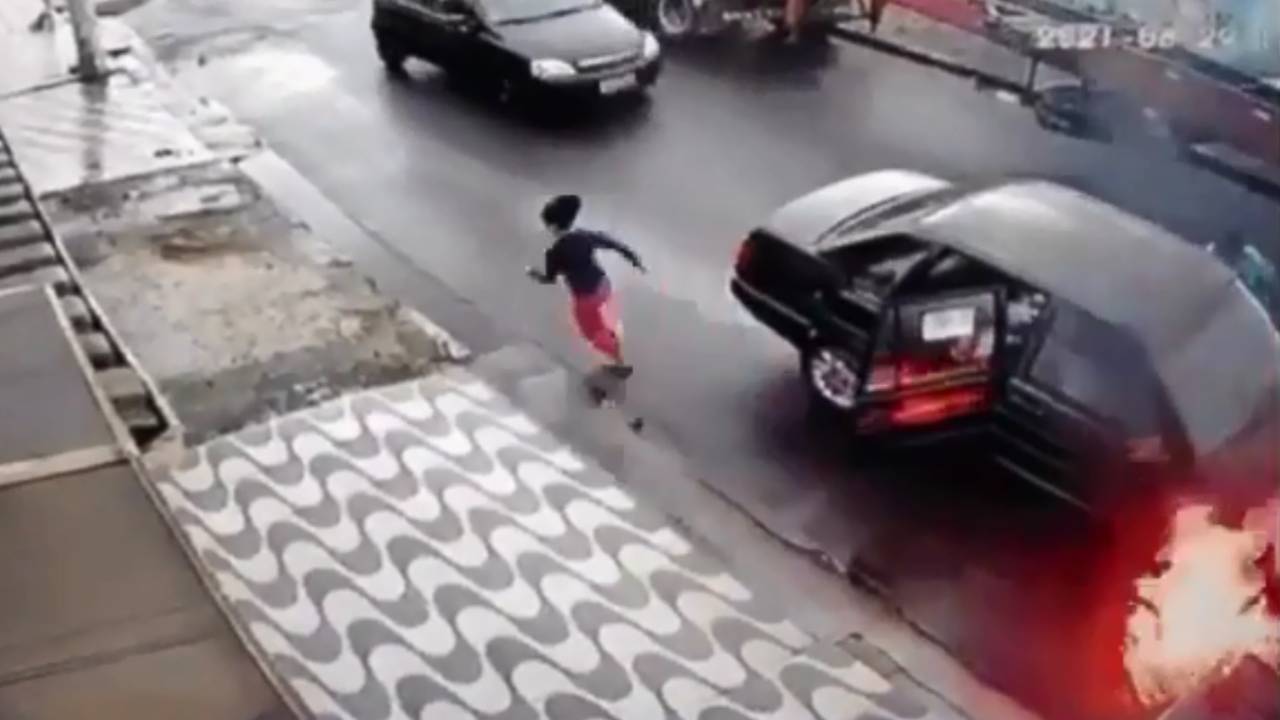 A imagem mostra um carro em chamas no meio de rua. Ao lado dele, há uma criança correndo que acabou de sair da porta de trás.