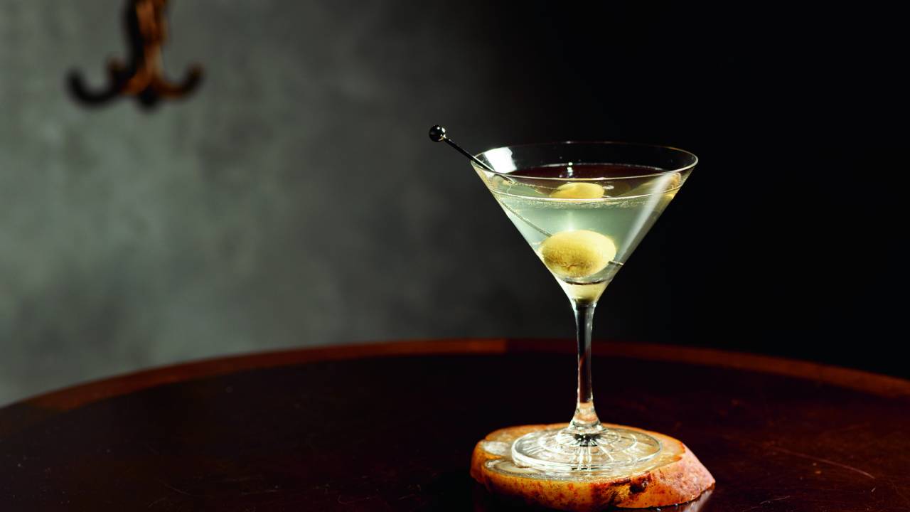 Em uma taça de martíni sobre mesa escura, drinque transparente com azeitona verde dentro.
