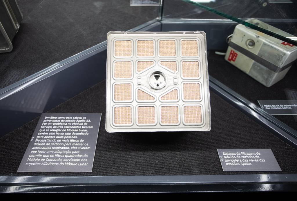 Filtro que que salvou a vida de tripulantes da Apollo 13 aparece dentro de vidro com quadradinhos amarelados em um quadrado de prata.