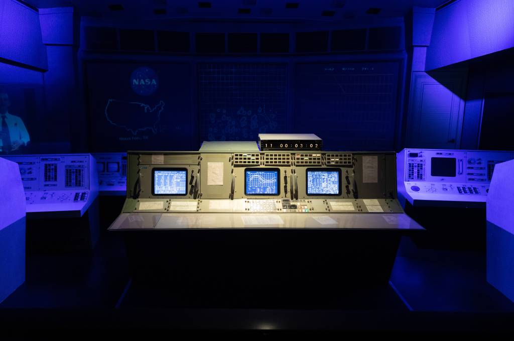 Painel de controle de missão espacial aparece iluminado em local escuro e azulado.