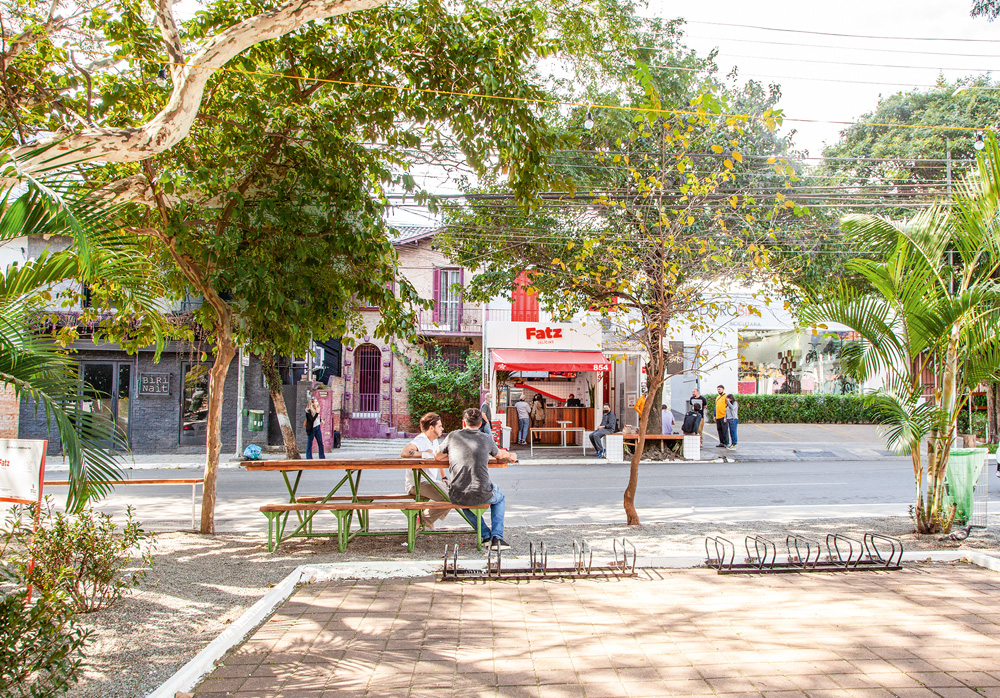 Foto tirada de praça em pinheiros da fachada da lanchonete Fatz Delícias do outro lado da rua.