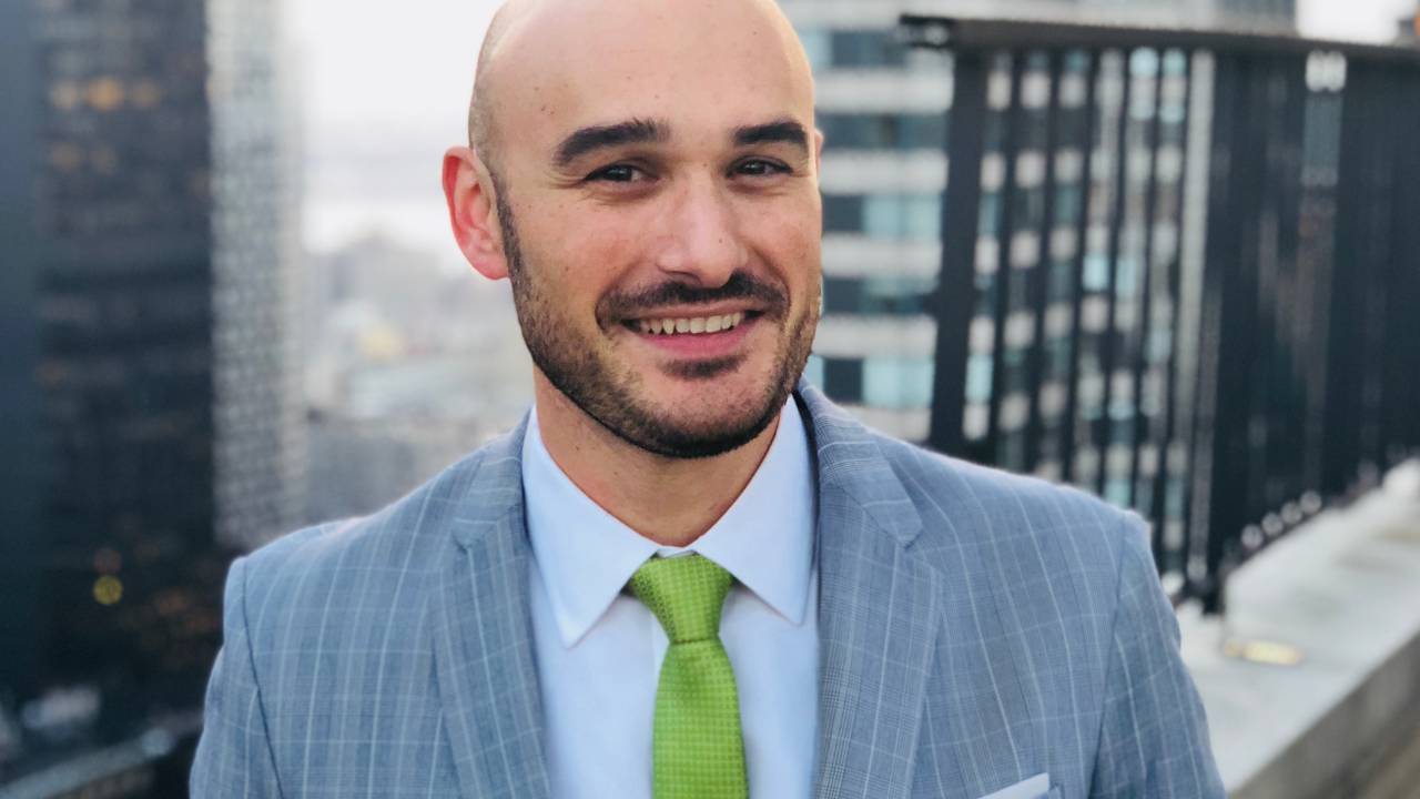 Gustavo Favaron, CEO do GRI Group, sorri para a câmera vestindo terno claro e gravata verde. Ele é careca, tem barba e posa em um terraço no alto de prédios.