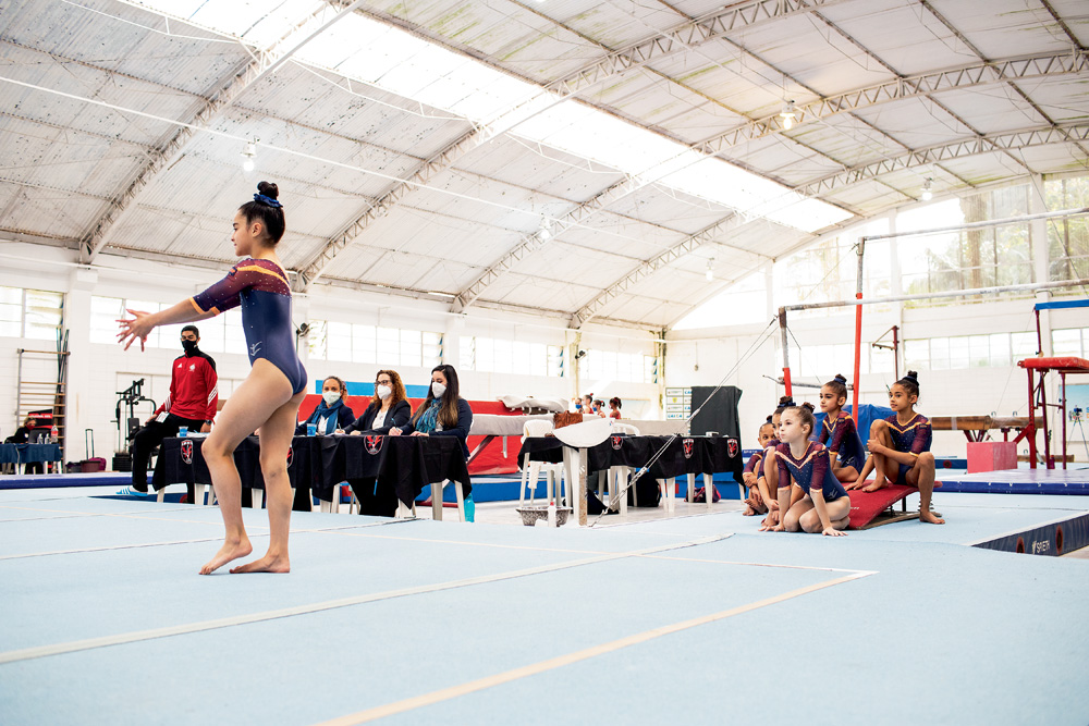 A imagem mostra o centro de ginástica de São Bernardo. Crianças estão observado uma ginasta preparando para fazer um movimento