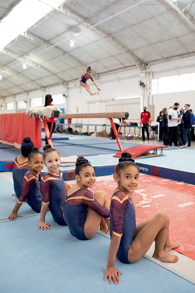 A imagem mostra cinco ginastas jovens sentadas a beira de um chão para a prática da ginástica.