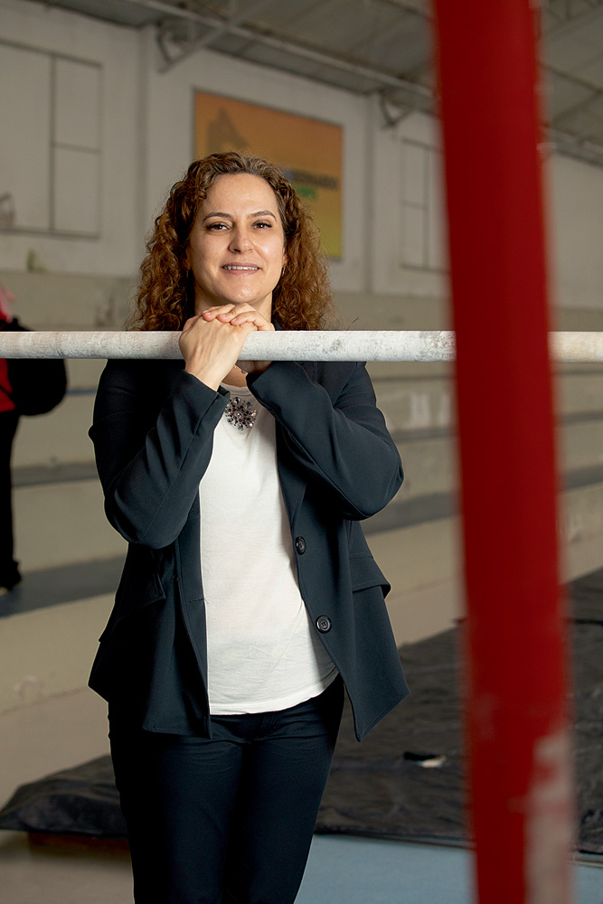 A imagem mostra Mônica, de pé, com as duas mãos apoiadas sobre uma barra de ginástica. Ela olha para a câmera sorrindo.