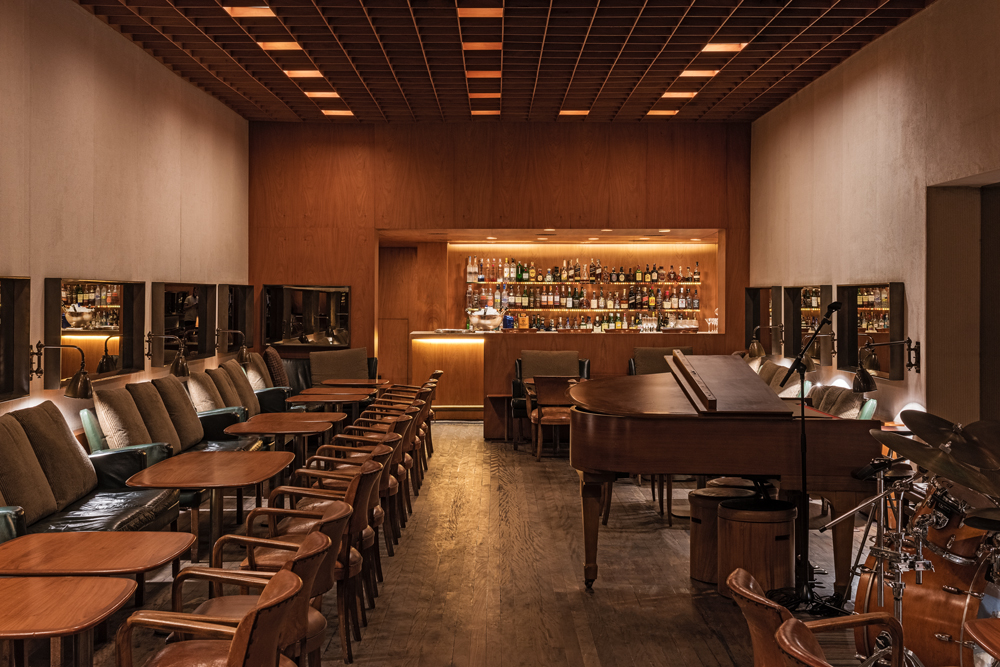 Salão do bar Baretto, com parede de madeira, balcão e prateleira de bebidas ao fundo. À direita, piano e bateria e à esquerda mesinhas e sofás.
