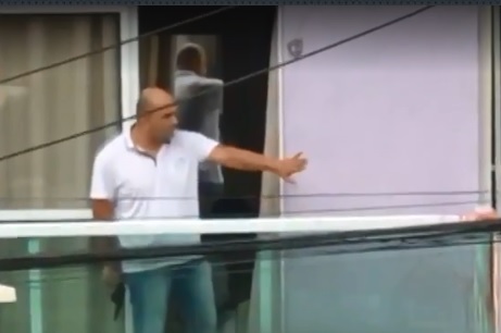 A imagem mostra um homem, em uma varanda, com uma arma na mão direita e a mão esquerda fazendo sinal para alguém se afastar.
