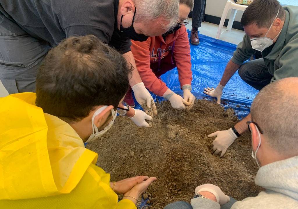 Pessoas de máscara aparecem agachadas mexendo em monte de terra.