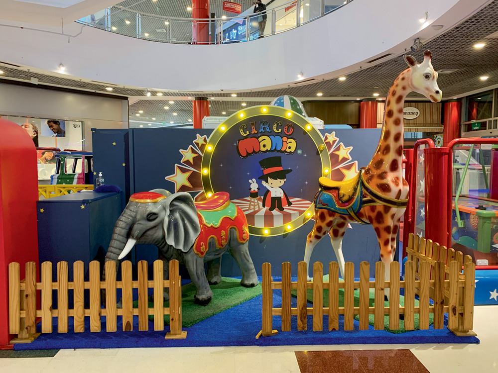 Um cercado dentro de um shopping tem a placa "Circo Mania Kids" com bonecos de elefante e girafa