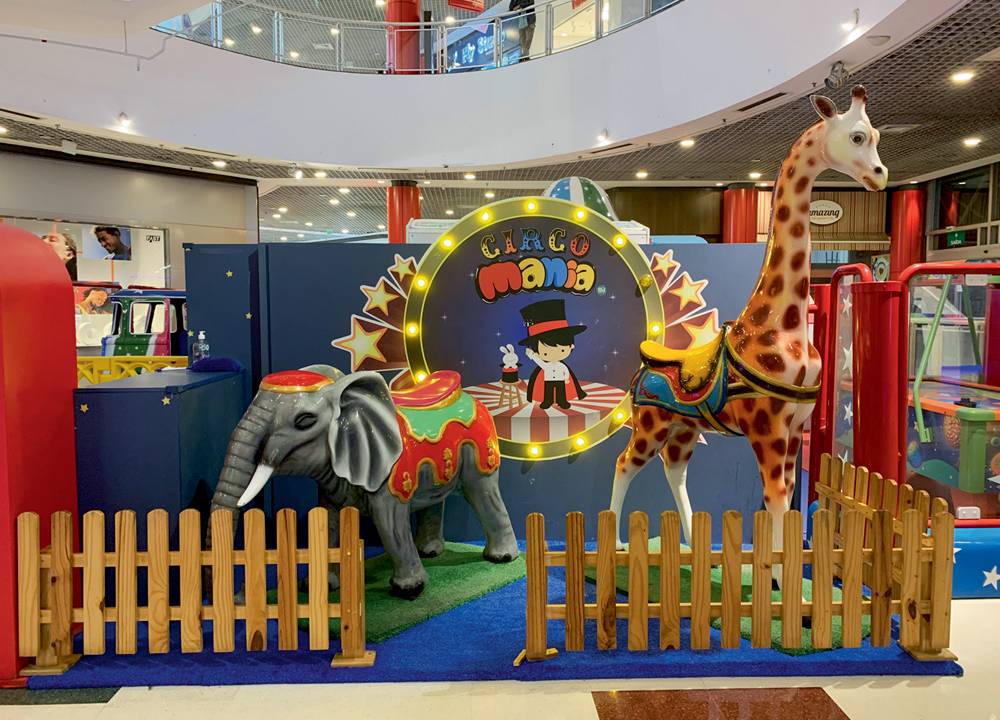 Um cercado dentro de um shopping tem a placa "Circo Mania Kids" com bonecos de elefante e girafa