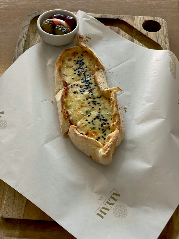 Alyah Sobre tábua de madeira clara, a esfirra alongada de queijo é servida junto de pequenos pedaços de picles, tomate e duas azeitonas pretas em um pequeno pote ao lado