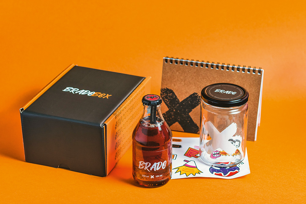 Caixa bradobox em fundo laranja ao lado de garrafinha de vidro com drinque, pote de vidro, cartela de adesivos e caderno pardo.