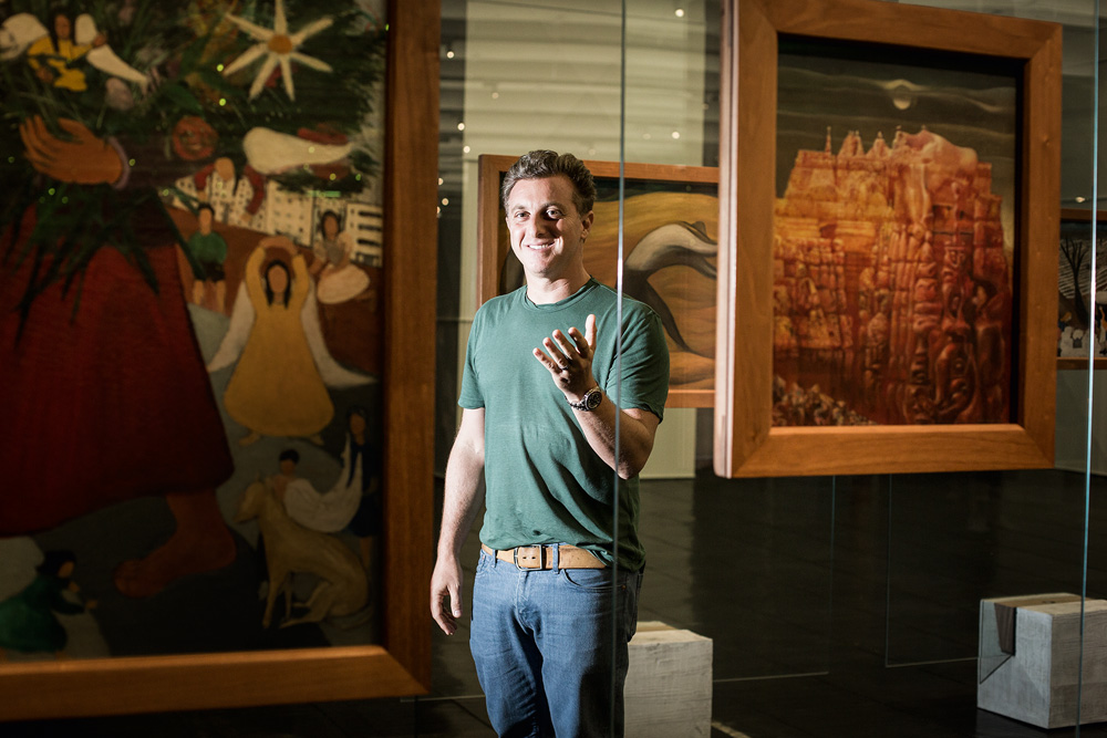 O apresentador Luciano Huck posa no Masp Museu de Arte de São Paulo. Veste camiseta verde e jeans, está de pé entre dois quadros e acena com uma das mãos erguida.