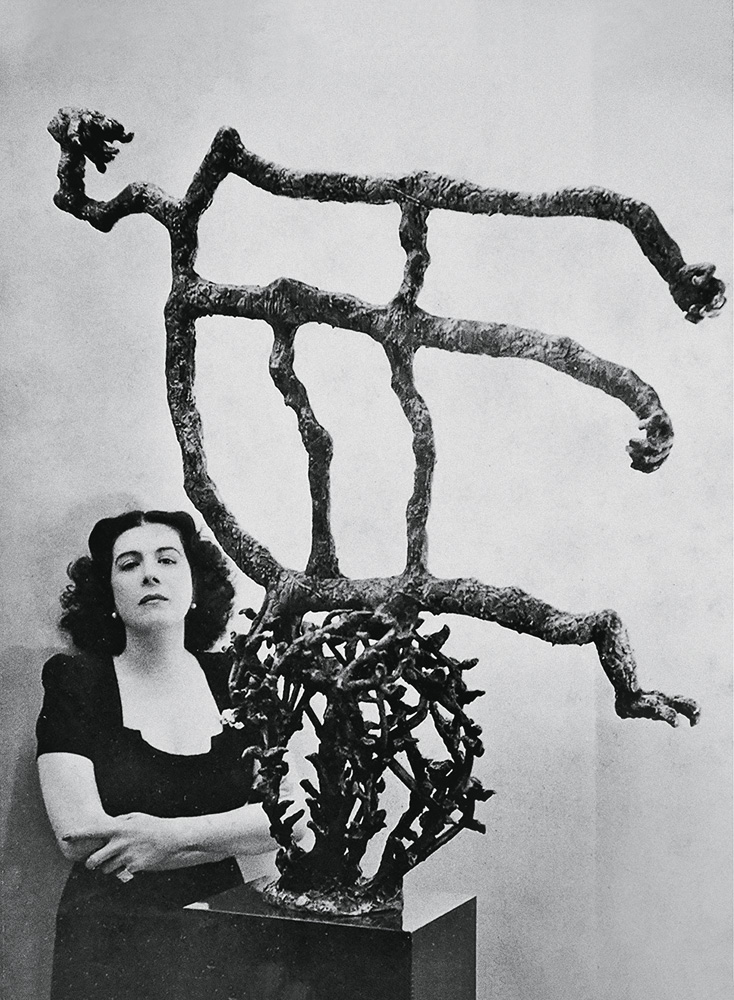A imagem mostra Maria Martins, de braços cruzados, ao lado de uma escultura surrealista feita por ela. São quatro galhos espinhentos, fazendo a forma de uma bola. Em cima deles, há outros galhos mais grossos e sem espinhos e sem formato definido.