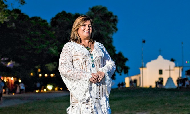 Martha Medeiros aparece de pé em gramado durante a noite. Ao fundo, uma casa branca. Ela veste vestido inteiramente branco e tem cabelos castanhos claros. Posa segurando as mãos uma na outra.