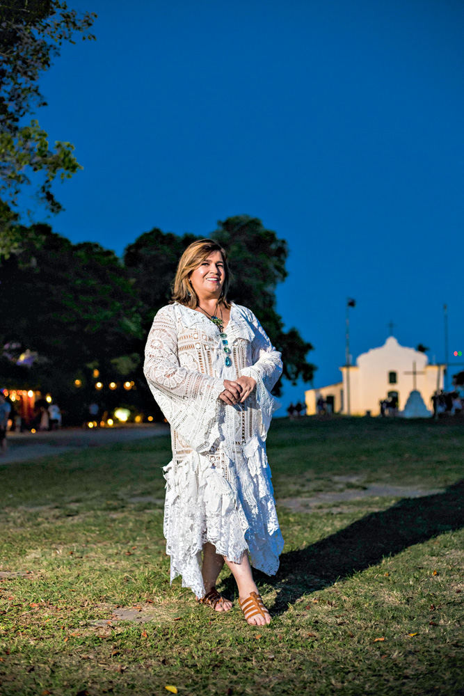 Martha Medeiros aparece de pé em gramado durante a noite. Ao fundo, uma casa branca. Ela veste vestido inteiramente branco e tem cabelos castanhos claros. Posa segurando as mãos uma na outra.