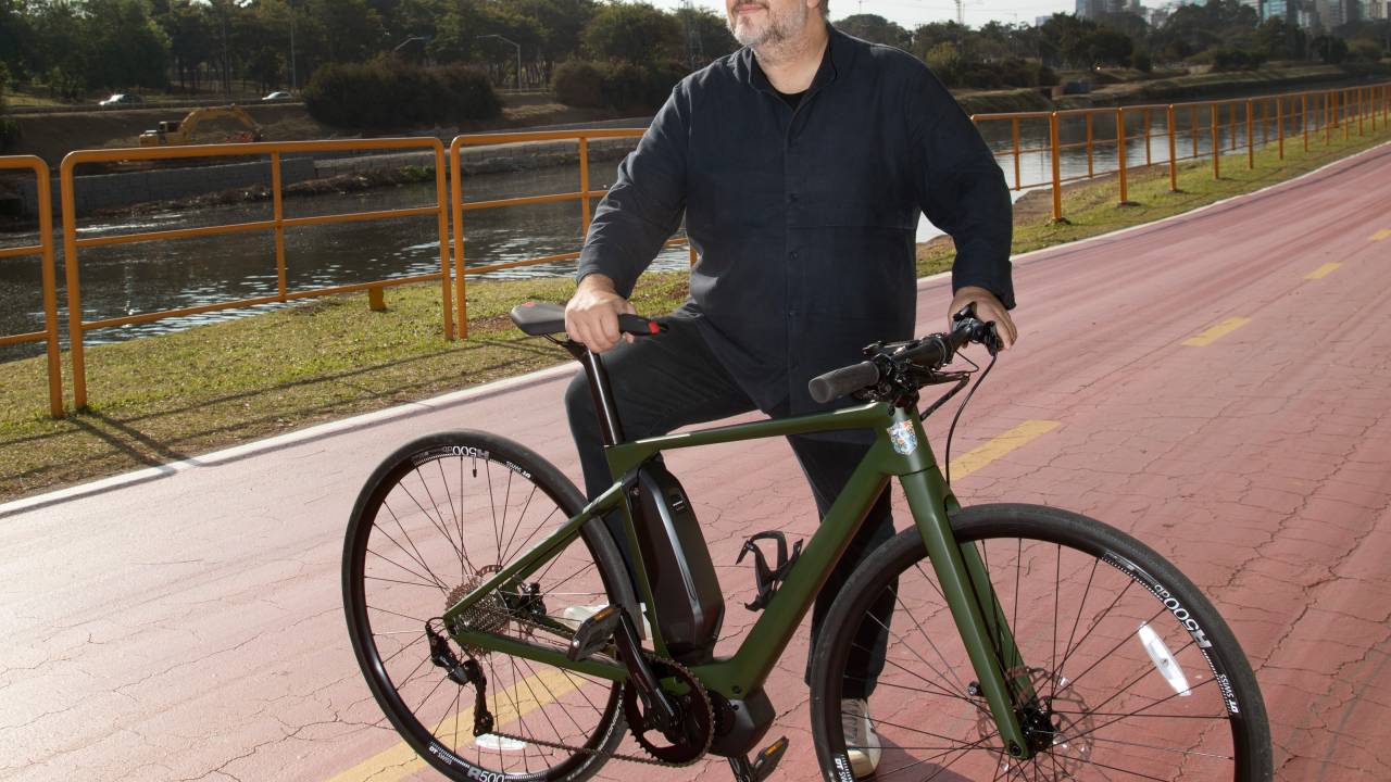 Michel Farah, da Farah Service, posa sentado na bike em uma ciclovia ao lado do Rio PInheiros. Olha para o horizonte e veste roupas pretas.