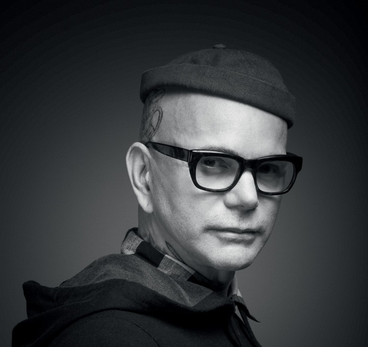 Hair stylist Neandro Ferreira encara a câmera em foto preto e branco. Usa óculos com armação preta grossa, usa gorro e jaqueta com capuz.