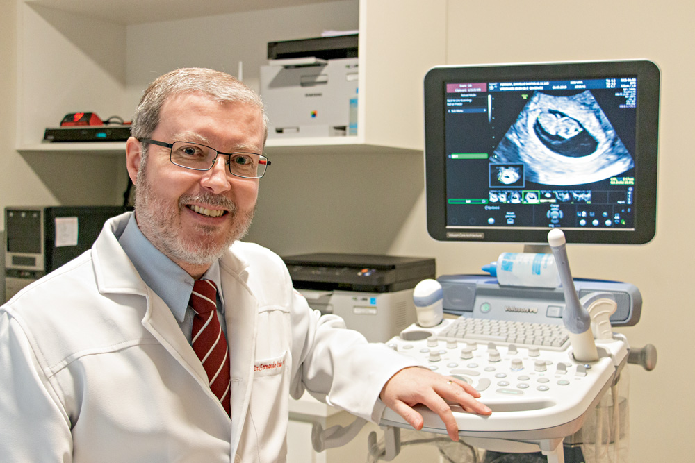 A imagem mostra Fernando, em um consultório médico, vestido com um jaleco branco. Ele está sentado, sorrindo para a foto e ao fundo há uma imagem de um ultrassom.