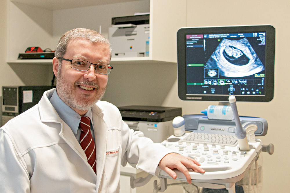Fernando Prado A imagem mostra Fernando, em um consultório médico, vestido com um jaleco branco. Ele está sentado, sorrindo para a foto e ao fundo há uma imagem de um ultrassom.