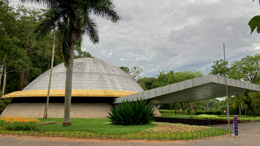 A imagem mostra uma foto do Planetário do Ibirapuera. É um domo, cinza, com uma entrada coberta por um telhado cinza.