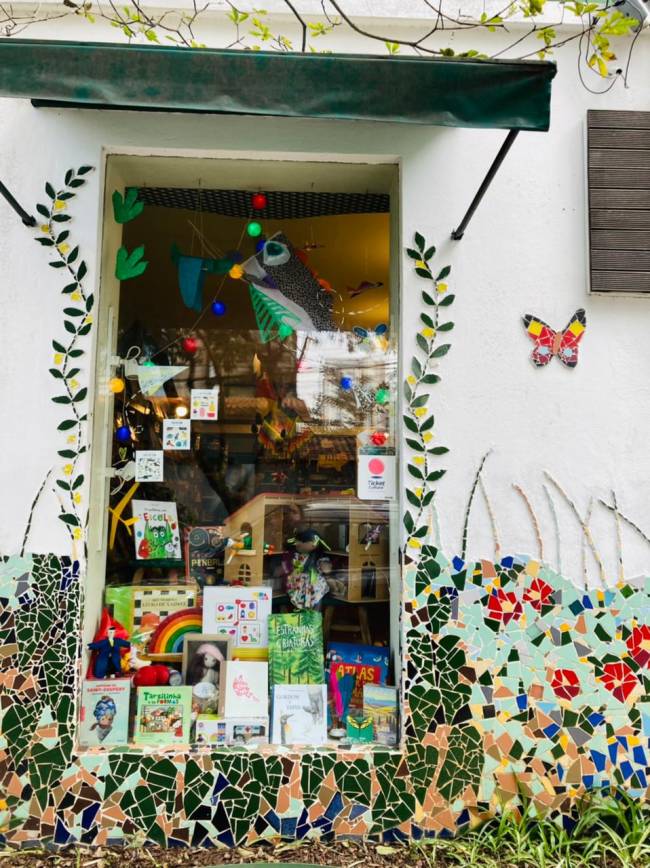 Foto exibe fachada da Livraria PanaPaná. decorada com pintura colorida de flores e vitrine de vidro com livros expostos.