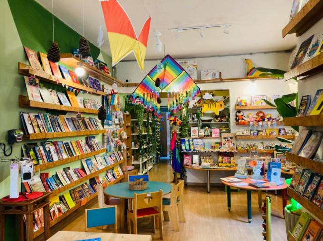 Foto exibe interior da Livraria PanaPaná, com decoração de papel no teto e prateleiras de madeira no canto esquerdo, com livros. No centro, mesinhas pequenas espalhadas.