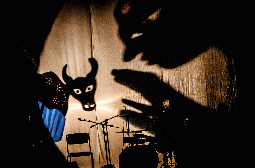 Em um teatro de sombras, dois bois são formados com as mãos em um palco. Há instrumentos musicais também