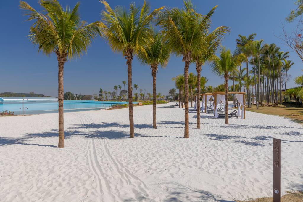 Foto mostra pedaço da praia artificial com ondas e areia em condomínio de luxo. Coqueiros aparecem em primeiro plano.