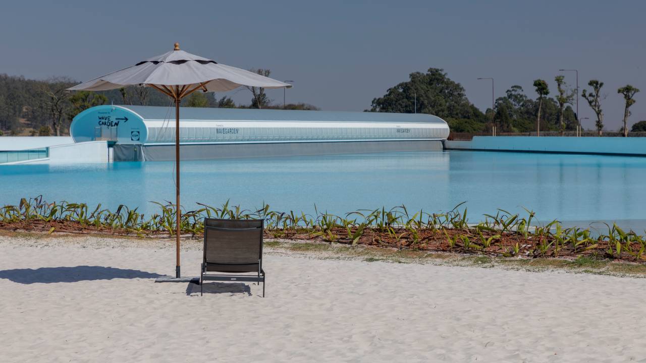 Foto mostra pedaço da praia artificial com ondas e areia em condomínio de luxo. Cadeira e guarda-sol aparecem em primeiro plano.