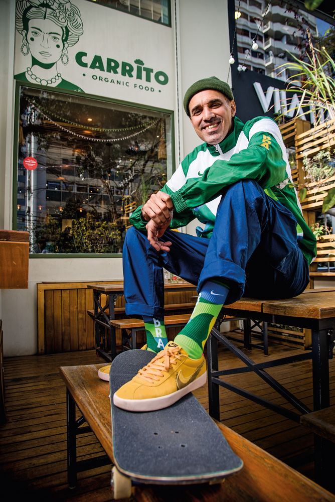 Homem posa com um pé apoiado em um skate, sentado em uma mesa. Ele está com uniforme do Brasil