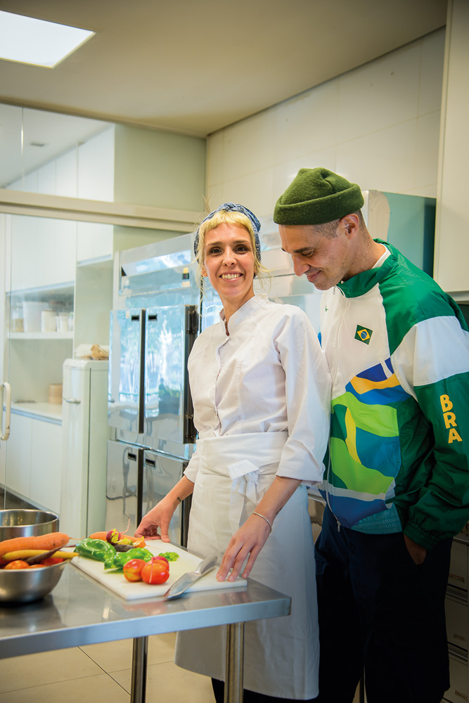 Uma mulher com roupas de cozinheira está em frente a uma bancada com comidas para picar. Um homem com roupas do Brasil está atrás dela. Os dois sorriem