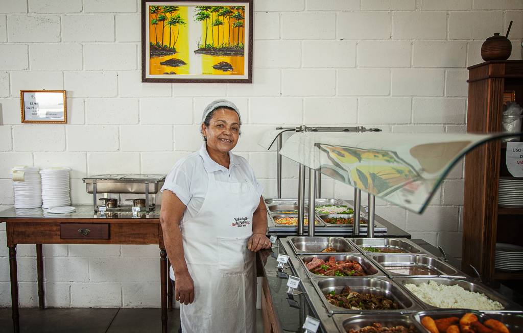 Marlene de avental e touca branca ao lado do bufê do Restaurante da Marlene.